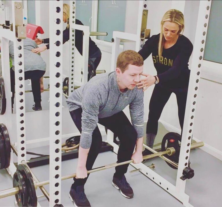 Professor Green lifting a barbell in a rack while a ROAR trainer checks his position, at ROAR Fitness London